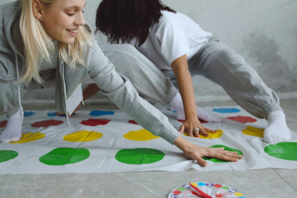 Two women playing a colorful Twister game indoors, having fun and laughing.