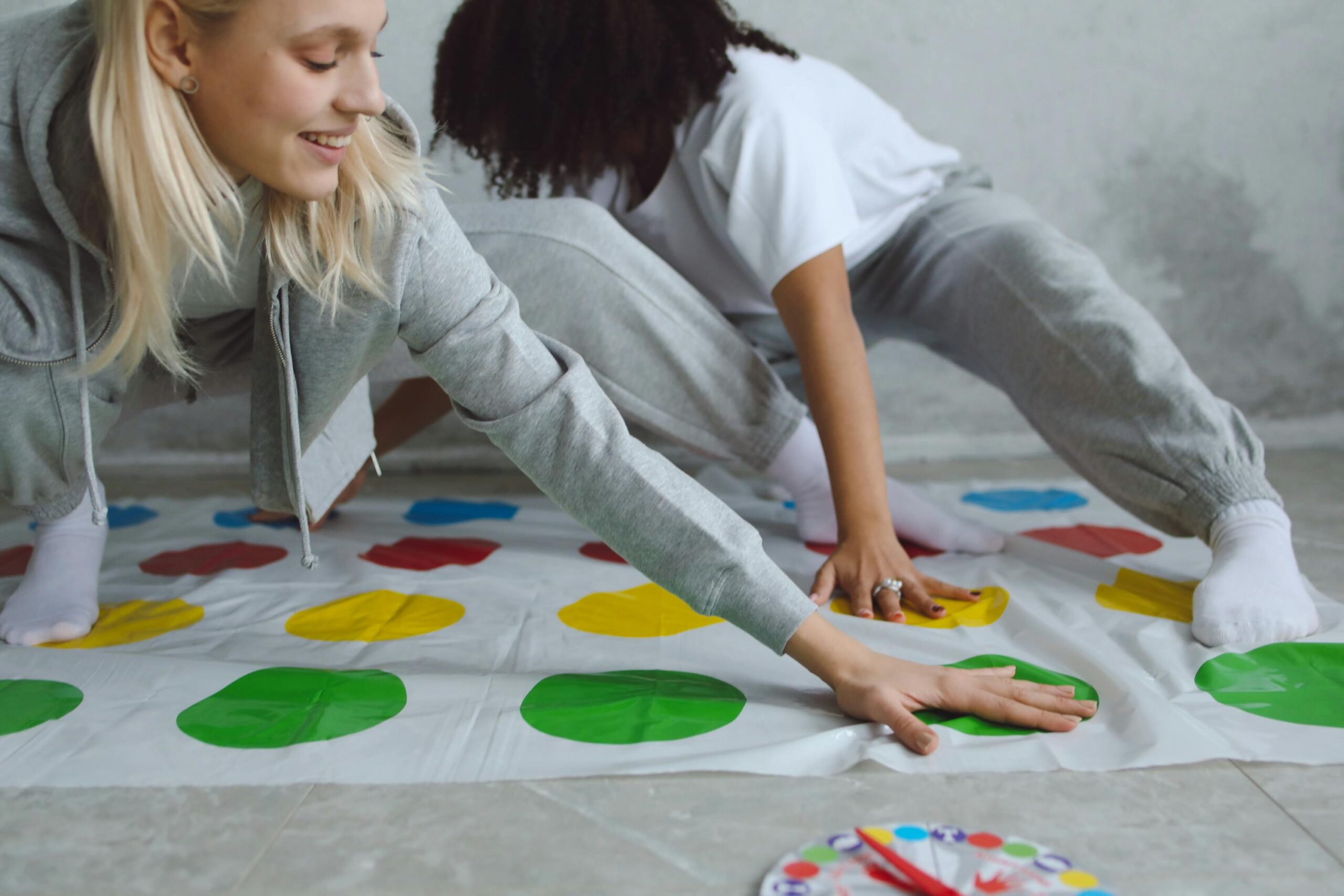 Two women playing a colorful Twister game indoors, having fun and laughing.