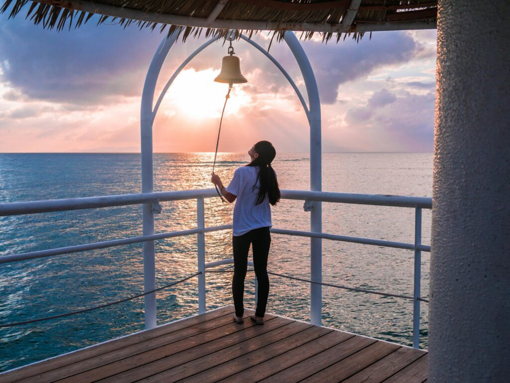 Woman standing on a seaside deck ringing a bell as the sun sets over the ocean.