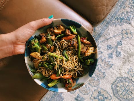 A close-up of a bowl of stir-fried noodles and vegetables being held by a person indoors.